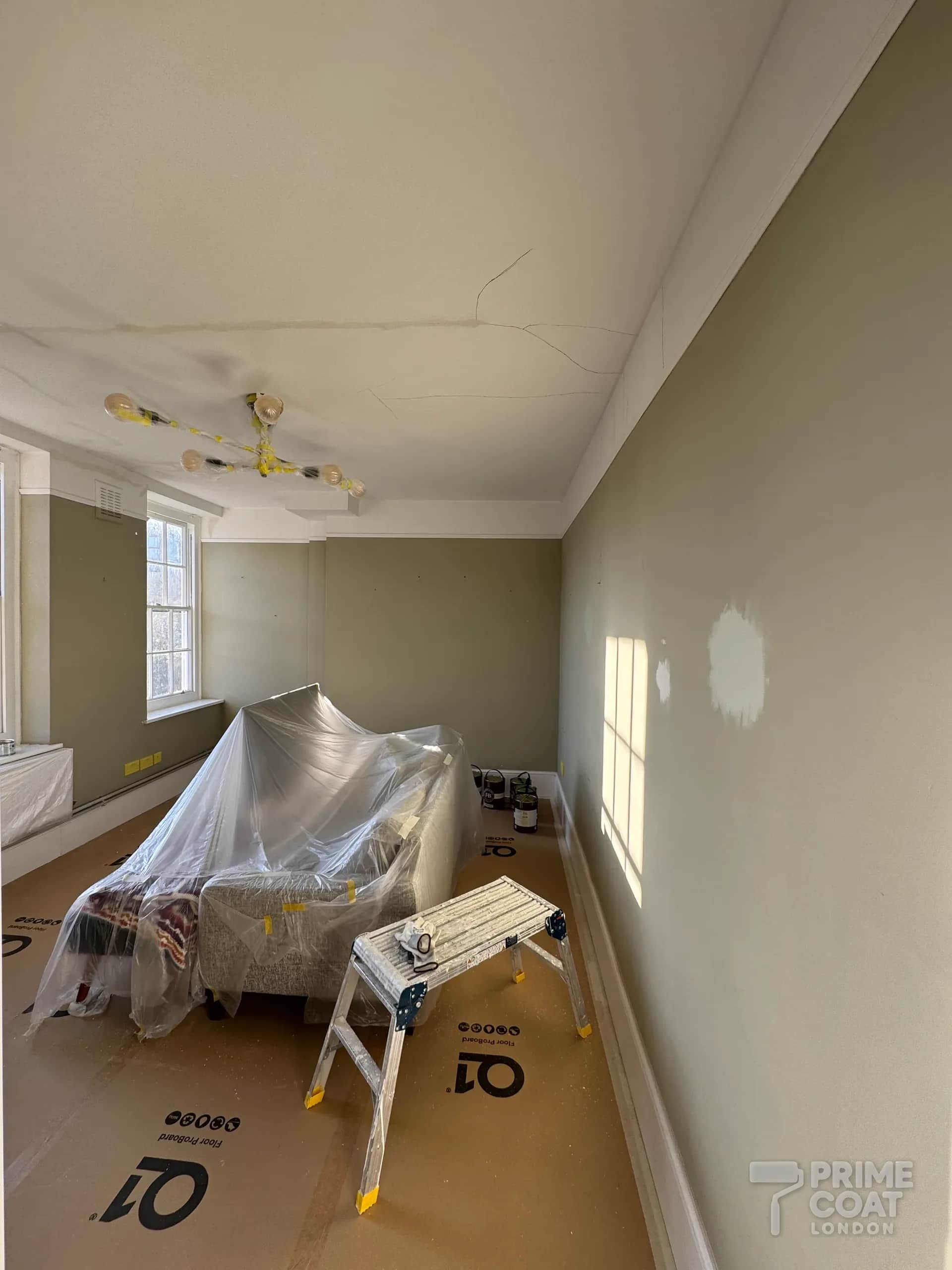 Battersea SW11 period flat living room before repainting — cracked ceiling with a water mark, patched walls, floor boarded and sofa wrapped