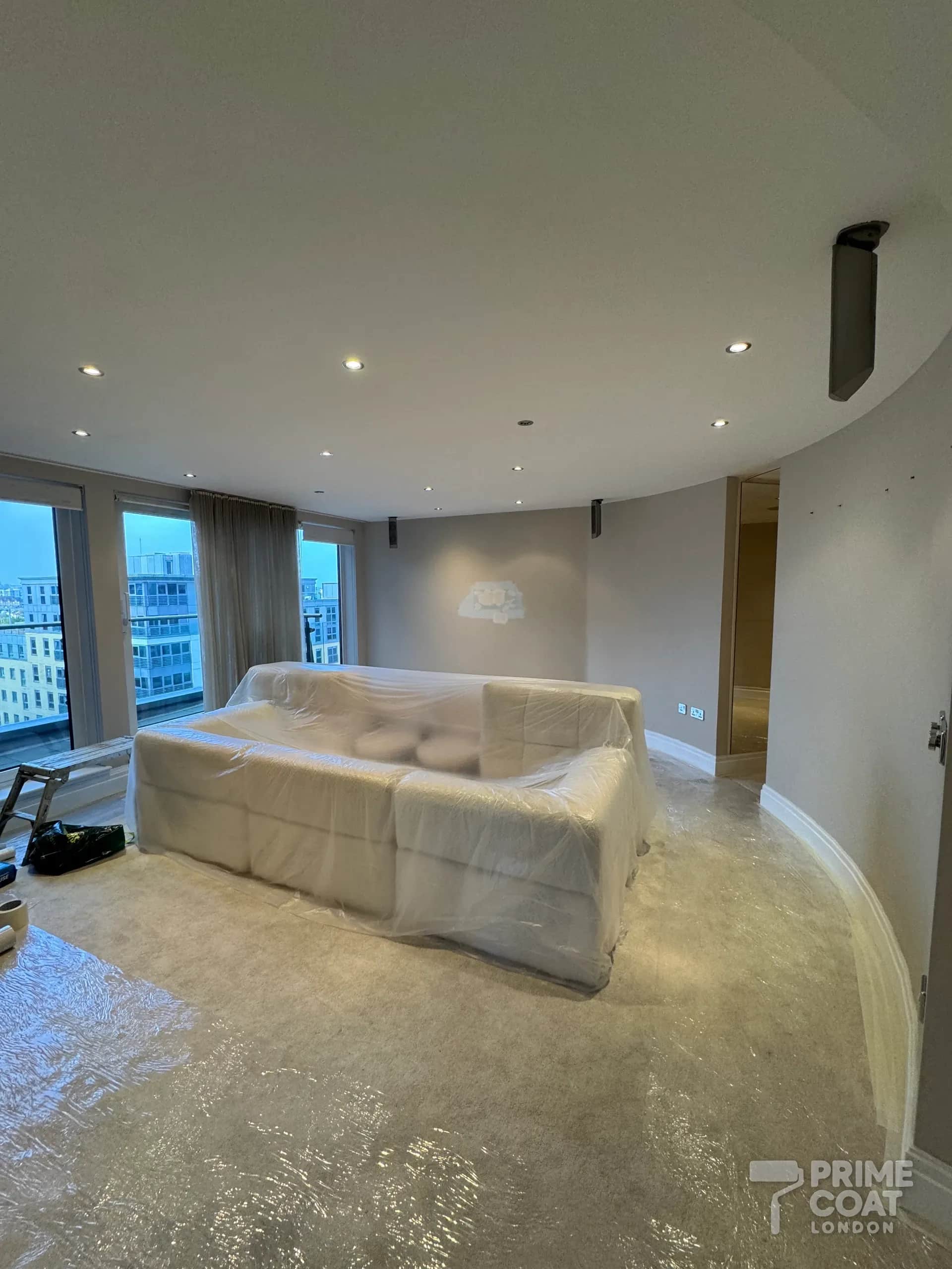 Chelsea Harbour SW10 living room during preparation — curved taupe walls with a filled repair, the sofa wrapped in plastic and the carpet sheeted