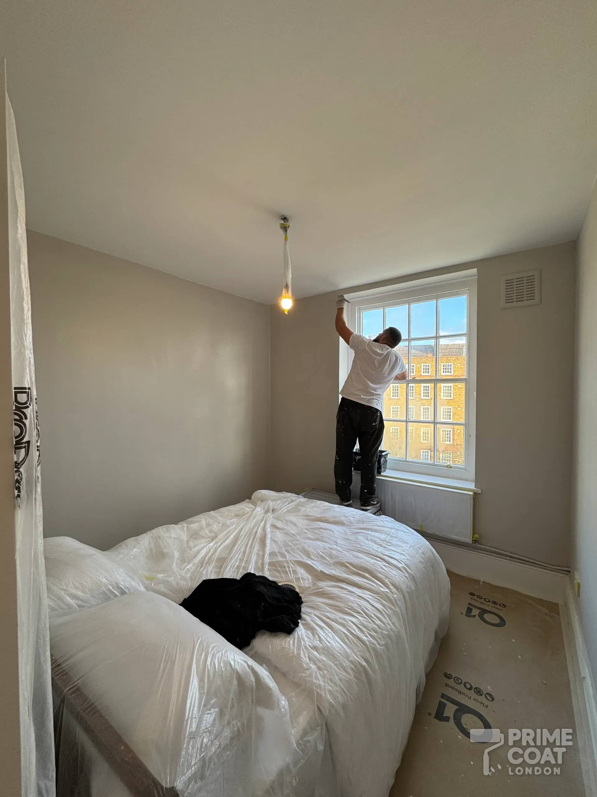 Decorator working at the sash window of a Battersea SW11 bedroom, the bed wrapped and Q1 boards protecting the floor