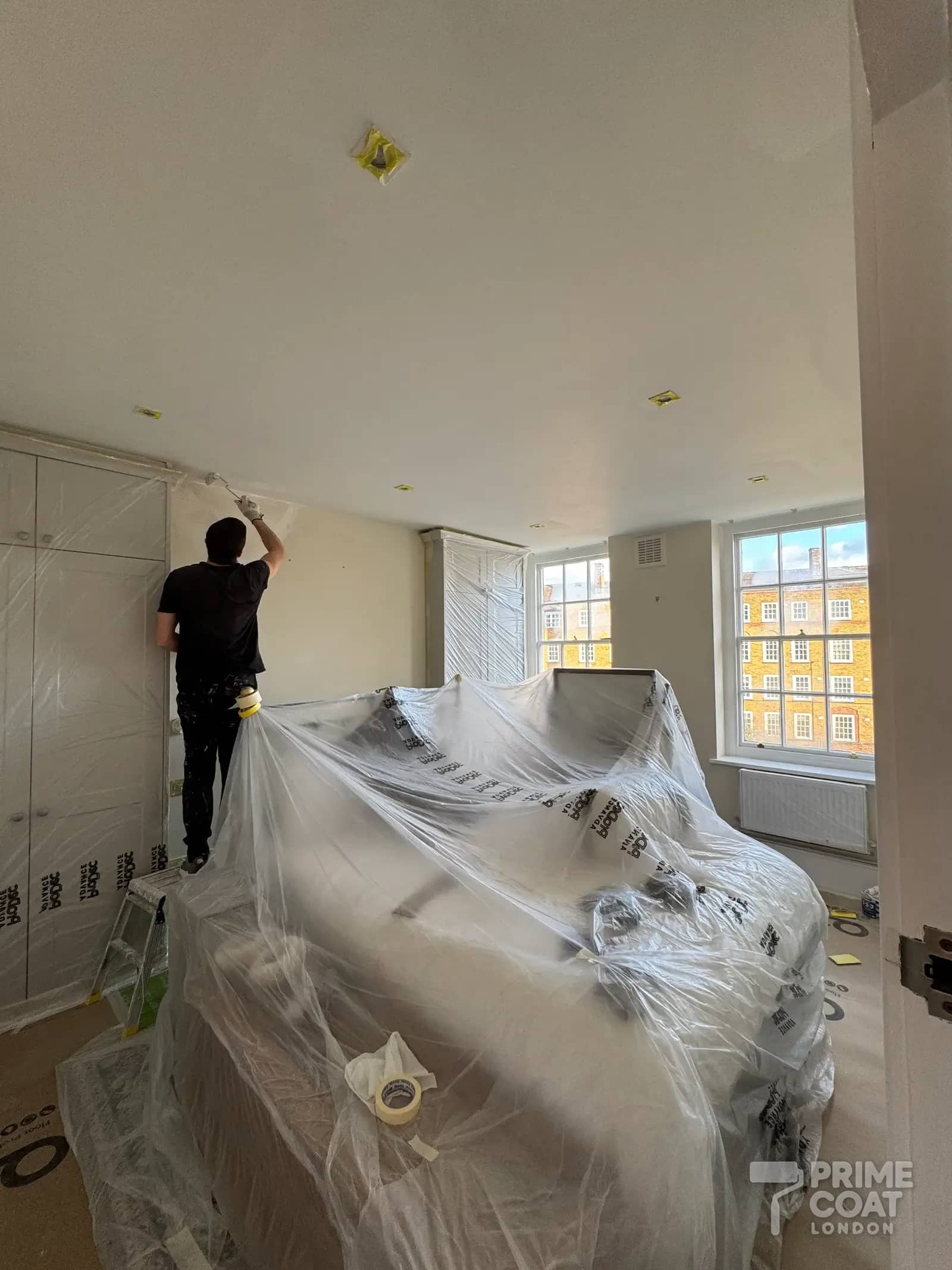Prime Coat decorator painting the master bedroom of a Battersea SW11 flat, with the bed and fitted wardrobes sheeted in plastic