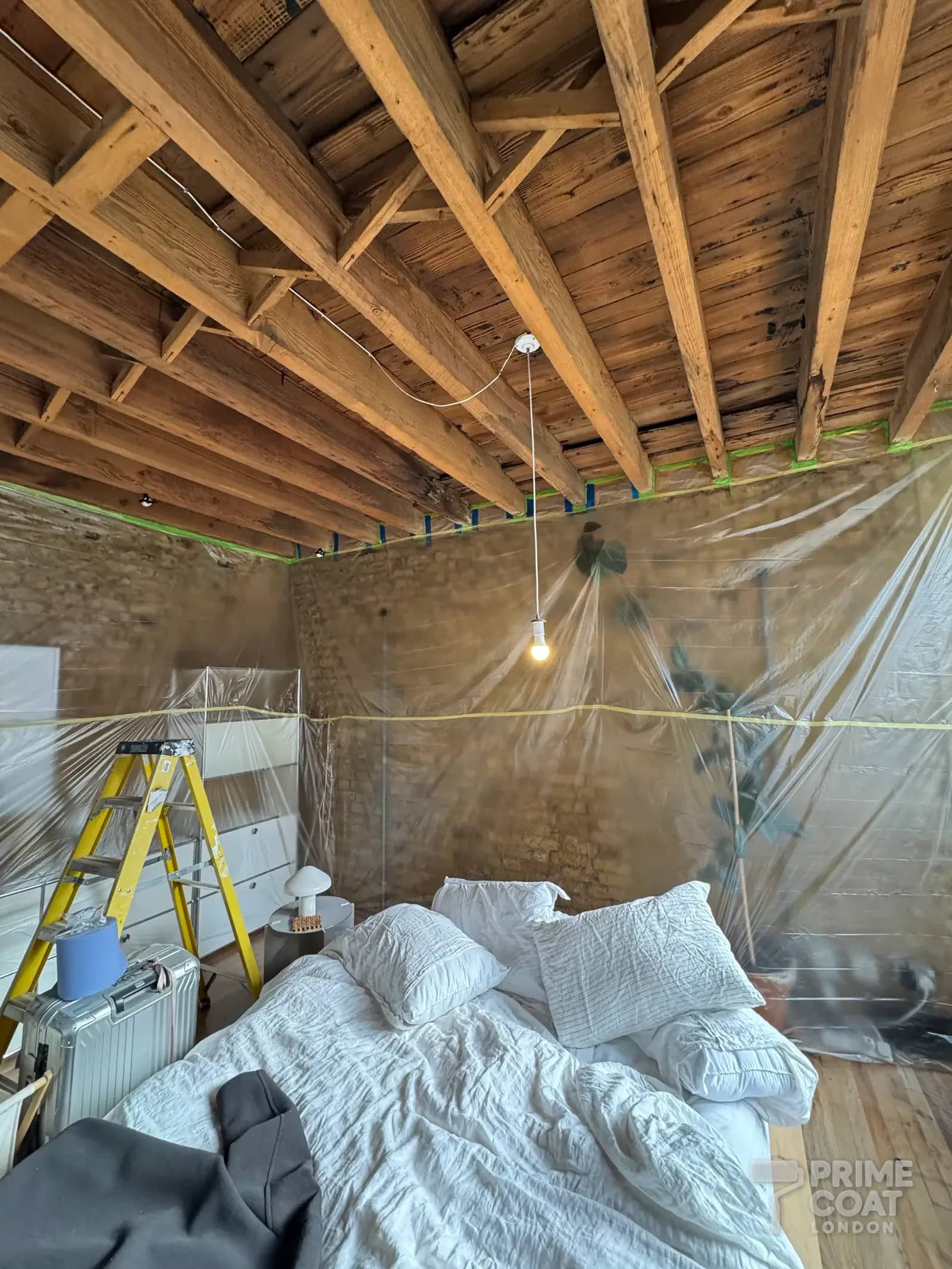 Bedroom in a Shoreditch E2 loft before painting — brick walls and furniture sheeted in plastic, edges taped beneath the raw timber ceiling