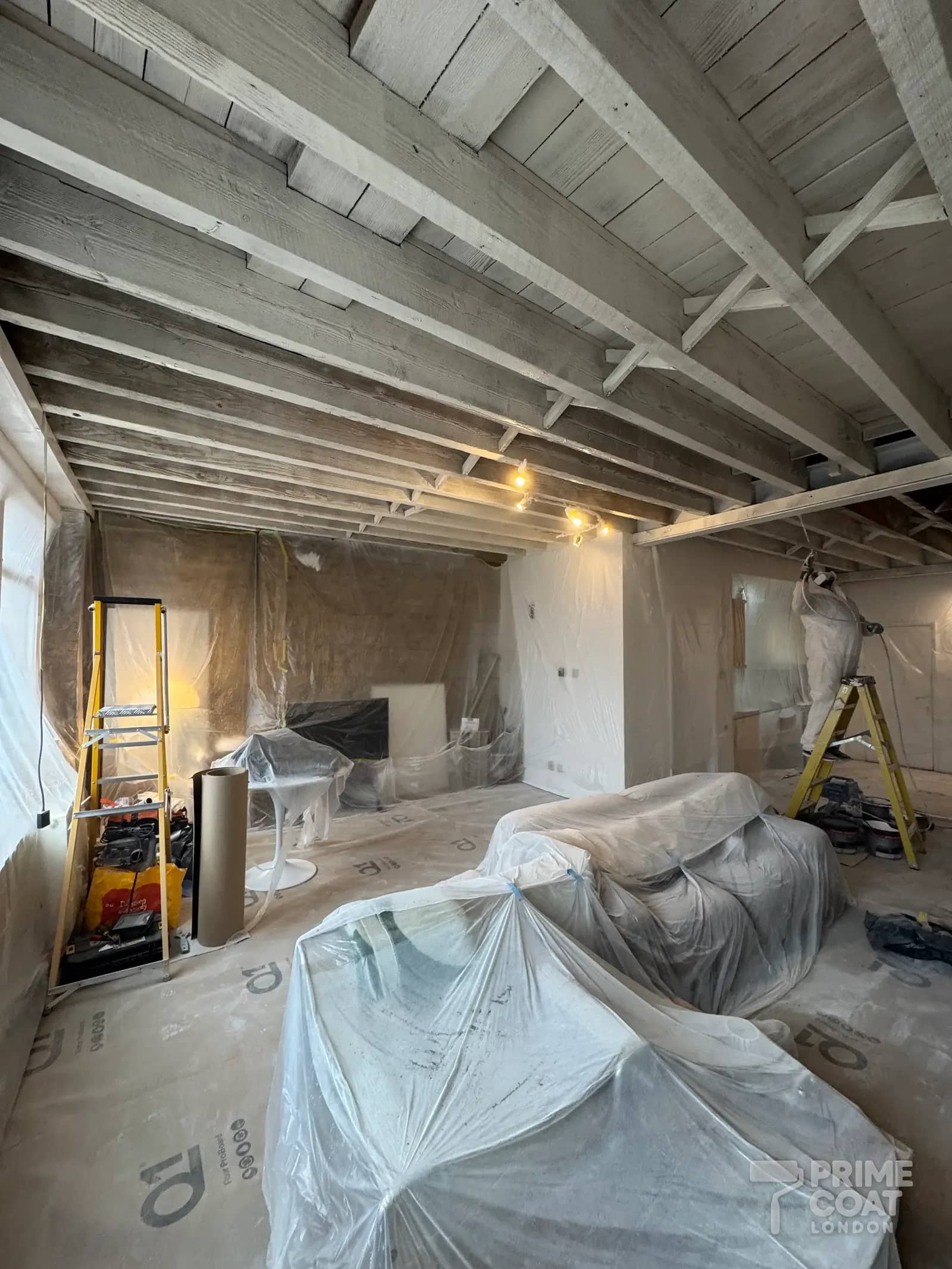 Decorator spraying the finish coat onto the timber ceiling of a Shoreditch E2 loft, with the room fully sheeted and Q1 boards on the floor