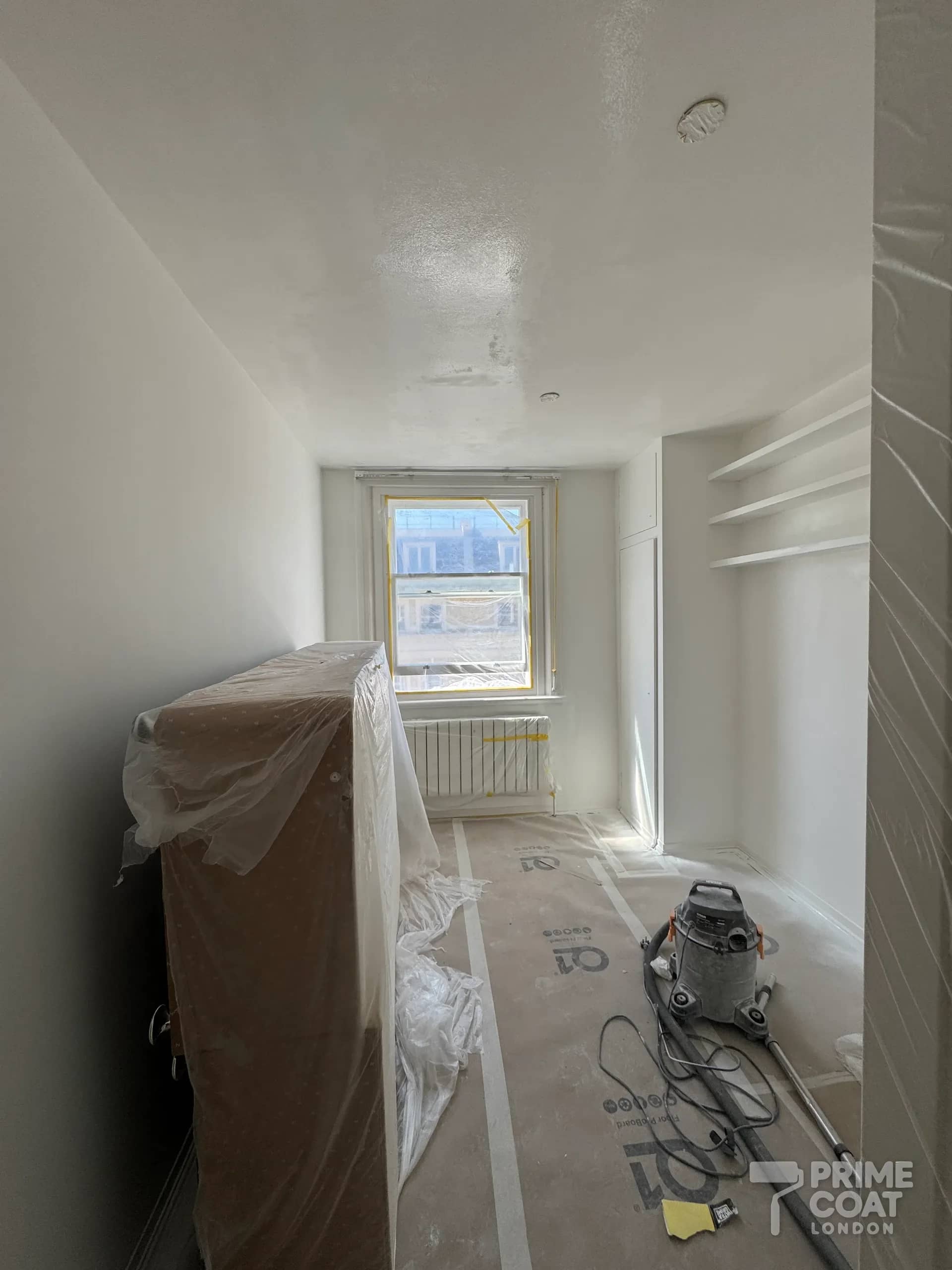 Bedroom in a Kensington W8 flat during spraying — sash window and wardrobe sheeted, ceiling and walls coated in white