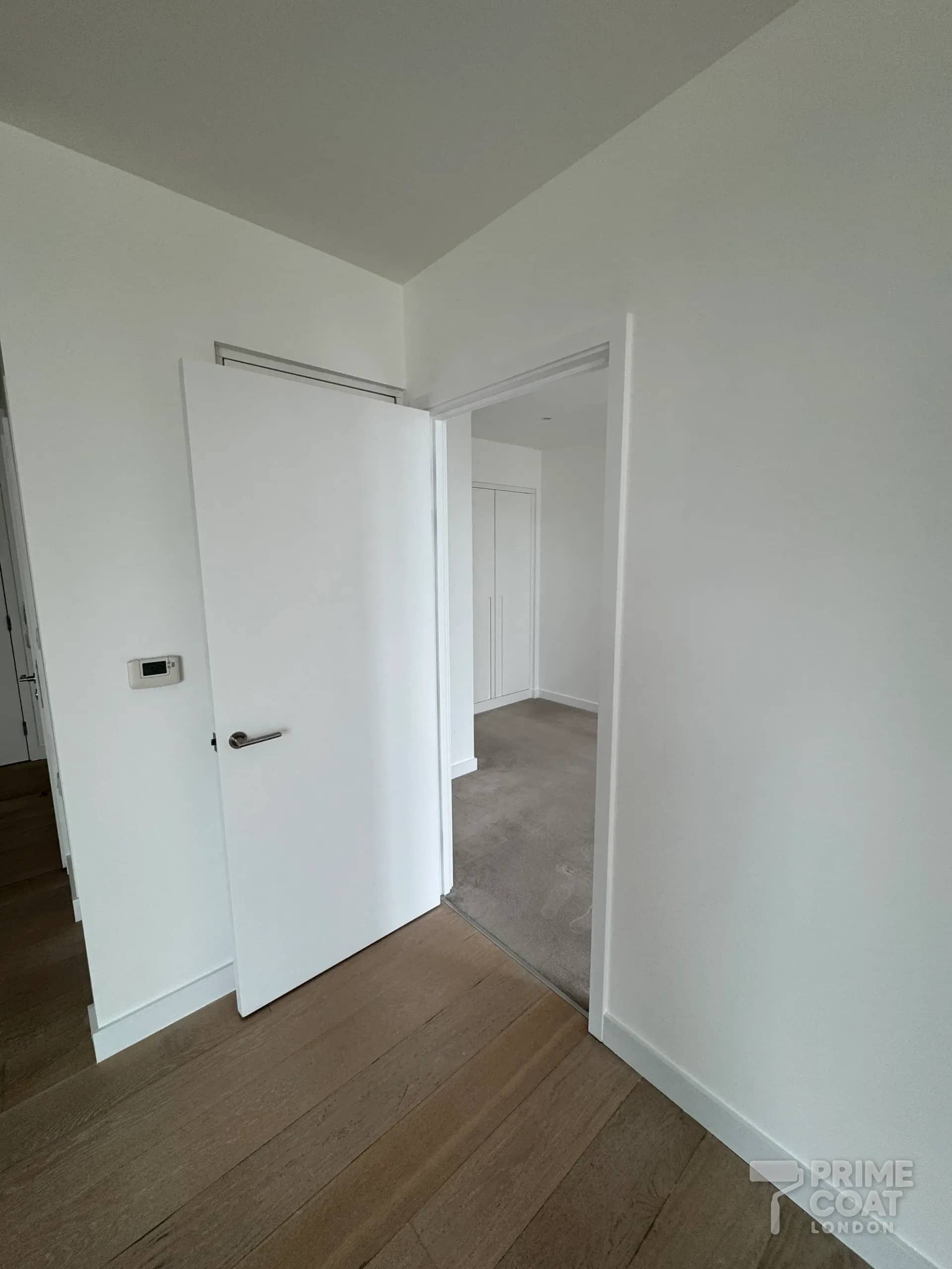 Hallway of a Pontoon Dock E16 apartment after repainting — white walls, a freshly painted white door and frame, and crisp white skirting on the oak floor, looking through to the bedroom